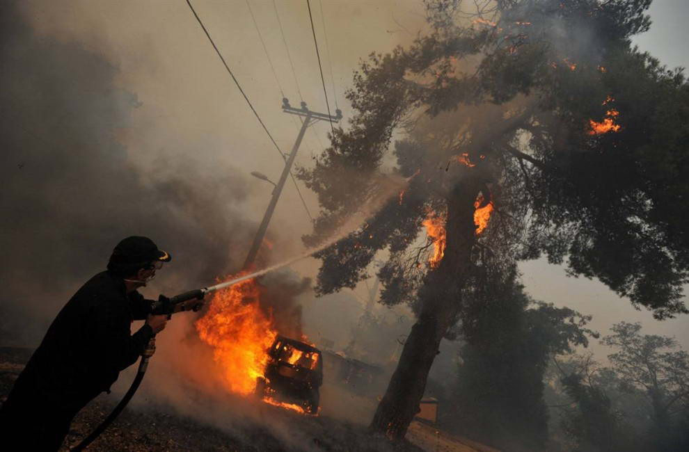 Пожарник тушит загоревшееся дерево на фоне горящего автомобиля недалеко от Агиос Стефанос. (Nikolas Giakoumidis/AP)